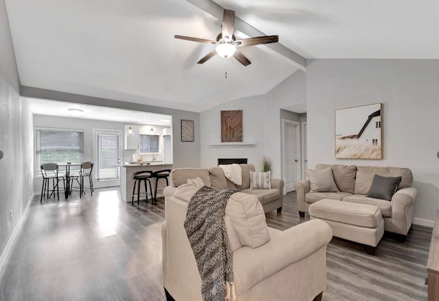 living area featuring a ceiling fan, wood finished floors, lofted ceiling with beams, baseboards, and a brick fireplace
