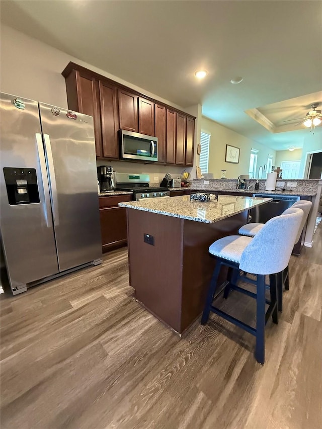 kitchen with wood finished floors, dark brown cabinetry, stainless steel appliances, light stone countertops, and a kitchen island
