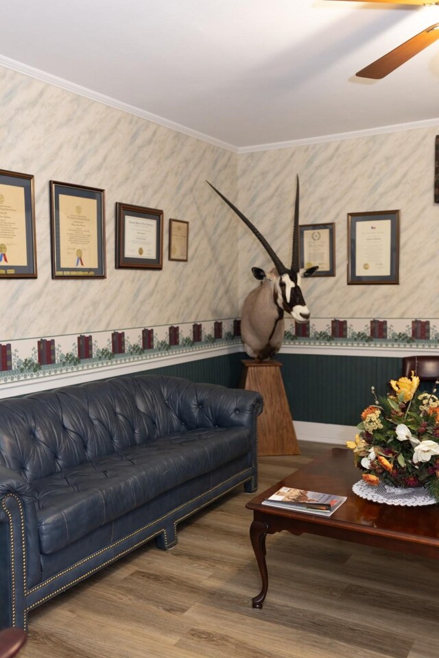 living area featuring wallpapered walls, wood finished floors, ornamental molding, and ceiling fan