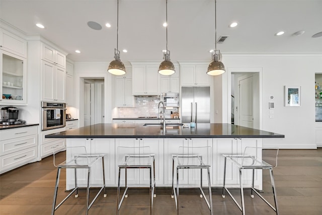 kitchen with dark countertops, a sink, built in appliances, ornamental molding, and white cabinetry