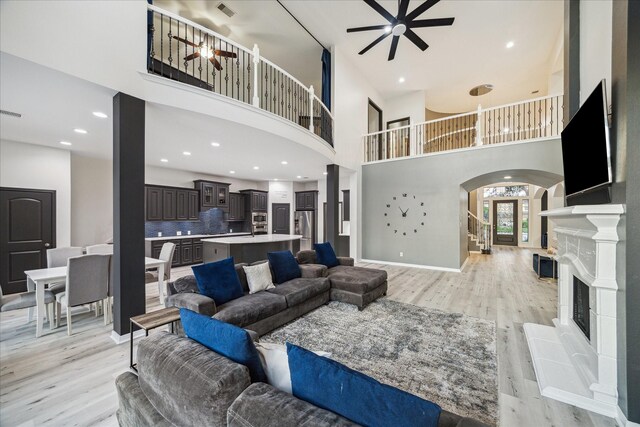living room featuring a high ceiling, a fireplace with raised hearth, arched walkways, a ceiling fan, and light wood-style flooring