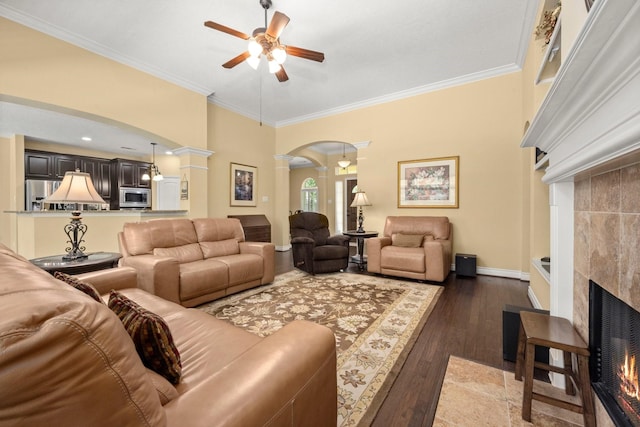 living room with ornamental molding, a tiled fireplace, ceiling fan, arched walkways, and ornate columns