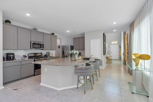 kitchen with tasteful backsplash, gray cabinetry, light stone counters, an island with sink, and appliances with stainless steel finishes