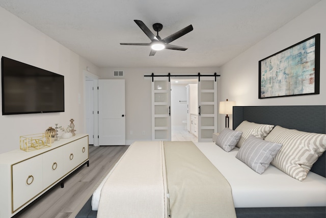 bedroom featuring visible vents, light wood-style floors, ensuite bathroom, a barn door, and a ceiling fan