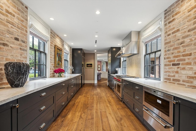 kitchen featuring baseboards, brick wall, exhaust hood, a warming drawer, and wood finished floors