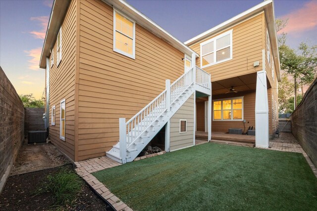 back of house at dusk with central AC, a fenced backyard, a yard, stairs, and ceiling fan