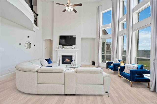 living room featuring a glass covered fireplace, light wood-style floors, baseboards, ceiling fan, and a high ceiling