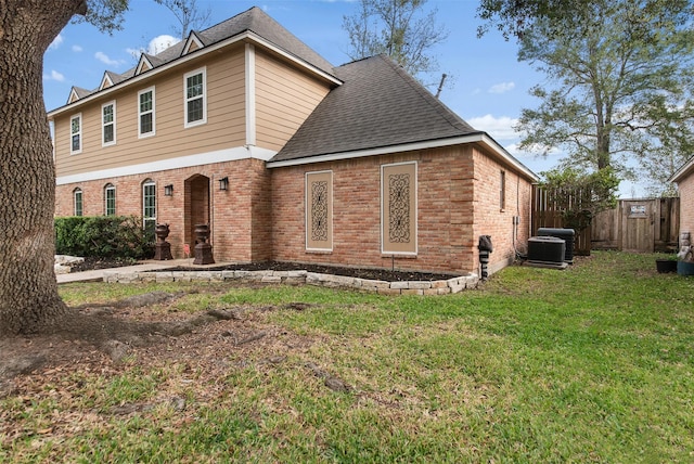 view of front facade featuring central AC unit, fence, a shingled roof, a front lawn, and brick siding