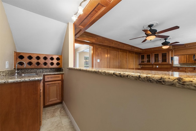 kitchen with a ceiling fan, a sink, brown cabinets, glass insert cabinets, and light stone countertops