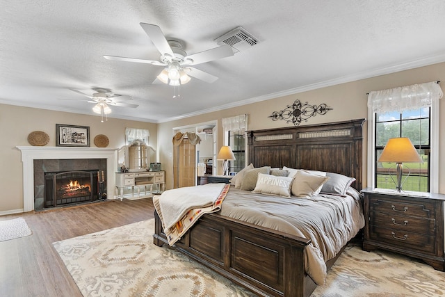 bedroom with a tiled fireplace, ornamental molding, light wood-type flooring, visible vents, and a textured ceiling