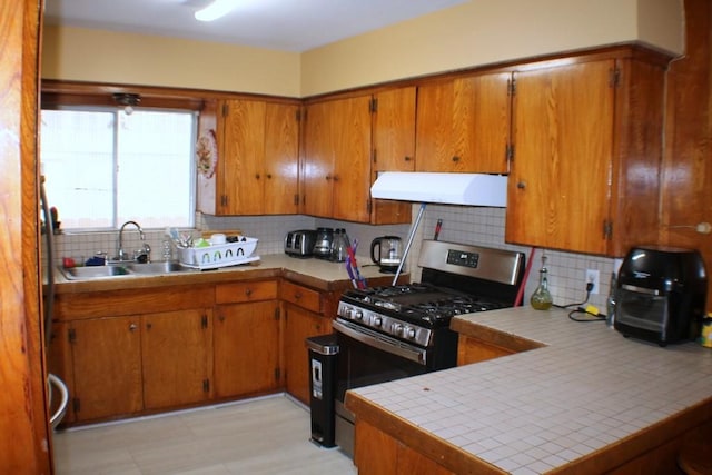 kitchen with decorative backsplash, range hood, stainless steel gas range oven, brown cabinets, and a sink