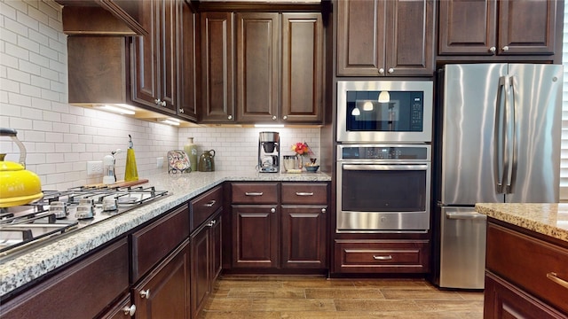 kitchen with wood finished floors, dark brown cabinetry, light stone counters, appliances with stainless steel finishes, and backsplash
