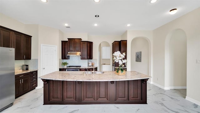 kitchen featuring a kitchen island with sink, a sink, marble finish floor, arched walkways, and freestanding refrigerator