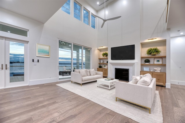living room with built in shelves, ceiling fan, a fireplace, baseboards, and light wood-type flooring