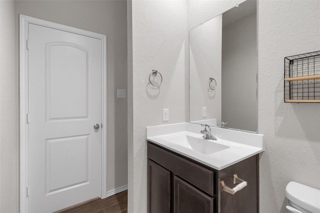 half bathroom with toilet, a textured wall, wood finished floors, and vanity