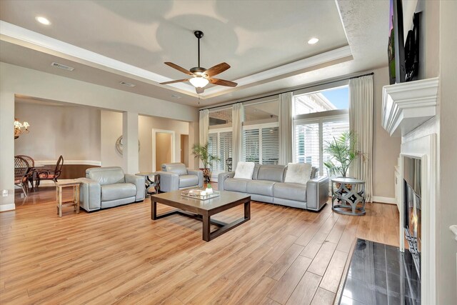 living area with visible vents, a fireplace with flush hearth, light wood-type flooring, ceiling fan with notable chandelier, and a tray ceiling