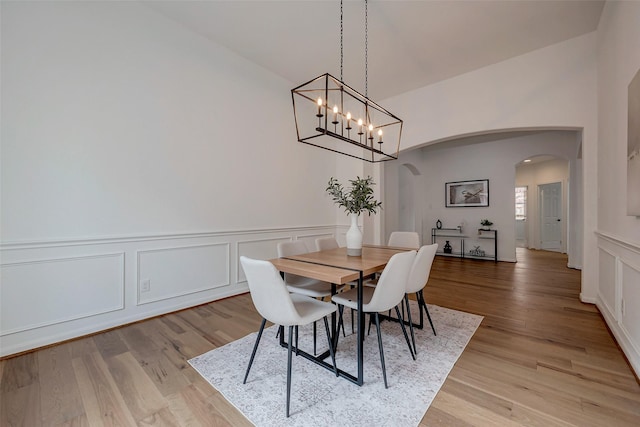 dining area featuring arched walkways, light wood-style flooring, a decorative wall, wainscoting, and a notable chandelier