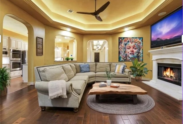 living room featuring visible vents, ceiling fan, a lit fireplace, a raised ceiling, and arched walkways