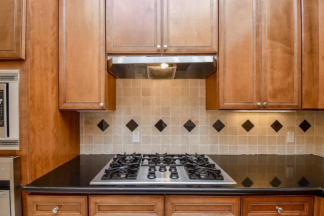 kitchen featuring brown cabinets, stainless steel appliances, under cabinet range hood, backsplash, and dark countertops