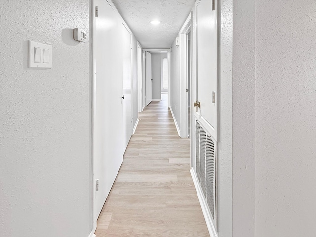 hallway featuring light wood-type flooring, visible vents, a textured wall, baseboards, and a textured ceiling