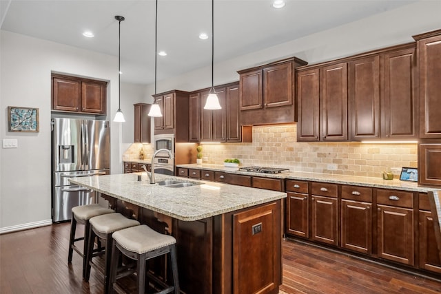 kitchen featuring a kitchen island with sink, a sink, stainless steel appliances, light stone counters, and dark wood finished floors