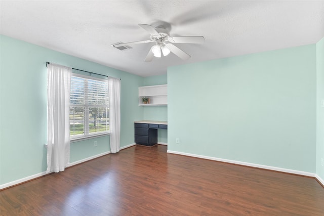 unfurnished room featuring ceiling fan, built in desk, baseboards, dark wood-type flooring, and visible vents