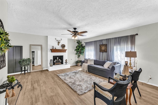 living room with a textured ceiling, baseboards, light wood finished floors, a brick fireplace, and a ceiling fan