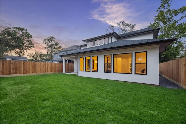 back of property at dusk with a lawn, stucco siding, a shingled roof, and a fenced backyard