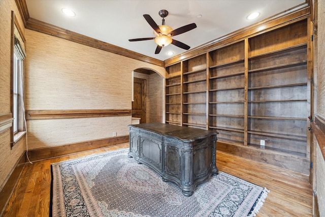 unfurnished office with crown molding, a ceiling fan, wood finished floors, built in shelves, and arched walkways