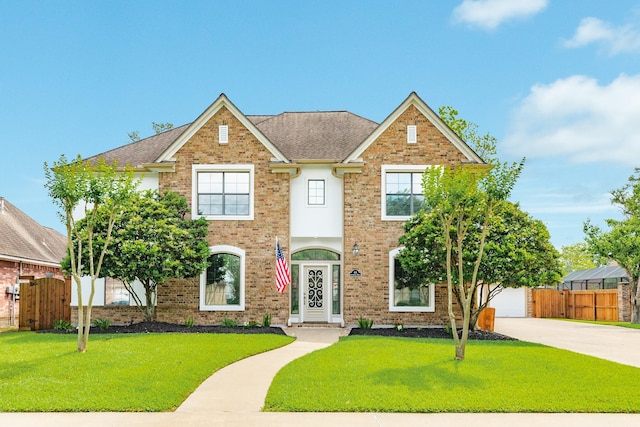 colonial-style house with a front yard, a shingled roof, brick siding, concrete driveway, and fence