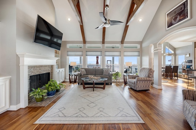 living room with wood finished floors, beamed ceiling, a brick fireplace, high vaulted ceiling, and a ceiling fan