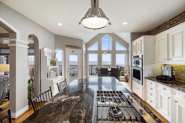 kitchen with dark stone countertops, arched walkways, a water view, double oven, and lofted ceiling