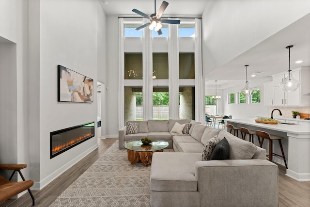 living room with a ceiling fan, a glass covered fireplace, light wood-style flooring, and a towering ceiling