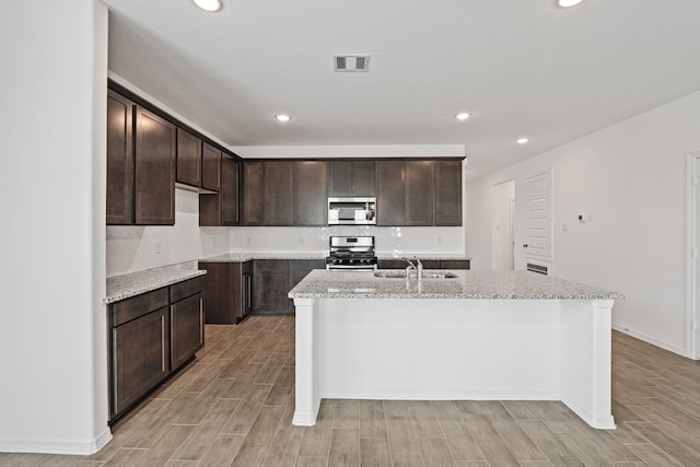 kitchen with a kitchen island with sink, a sink, stainless steel appliances, dark brown cabinetry, and wood finish floors