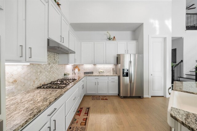 kitchen featuring light wood finished floors, under cabinet range hood, backsplash, white cabinetry, and appliances with stainless steel finishes