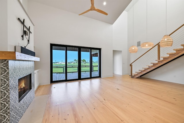 unfurnished living room with a tiled fireplace, stairway, light wood-type flooring, high vaulted ceiling, and a ceiling fan