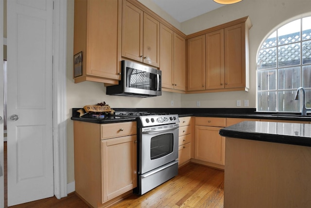 kitchen with light brown cabinetry, dark countertops, appliances with stainless steel finishes, a sink, and light wood finished floors