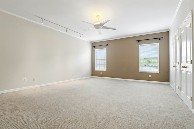empty room featuring rail lighting, a ceiling fan, baseboards, ornamental molding, and light colored carpet