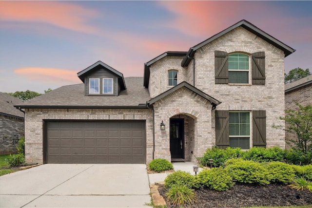french country style house with brick siding, an attached garage, roof with shingles, and concrete driveway