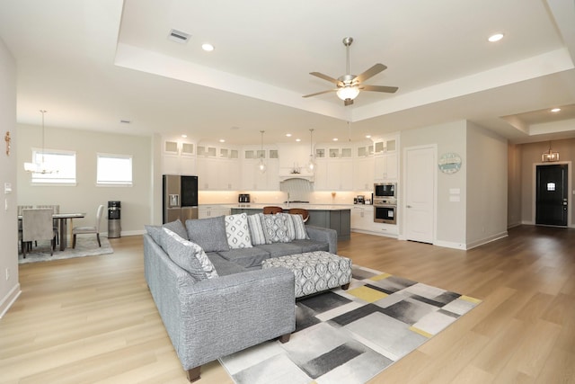 living room featuring visible vents, ceiling fan with notable chandelier, light wood finished floors, and a tray ceiling