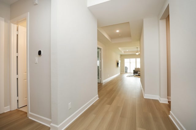 hallway featuring light wood-type flooring, recessed lighting, baseboards, and a tray ceiling