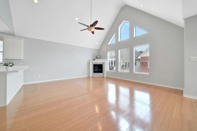 unfurnished living room featuring ceiling fan, high vaulted ceiling, a glass covered fireplace, light wood-type flooring, and baseboards