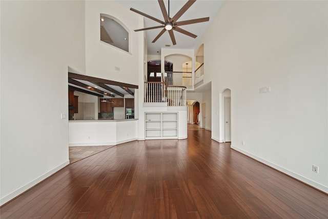 unfurnished living room featuring ceiling fan, arched walkways, wood finished floors, high vaulted ceiling, and baseboards