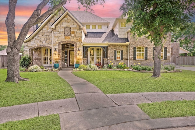 view of front of property with a front lawn, fence, stone siding, roof with shingles, and french doors