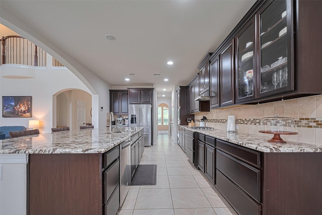 kitchen with arched walkways, appliances with stainless steel finishes, light tile patterned floors, light stone counters, and glass insert cabinets