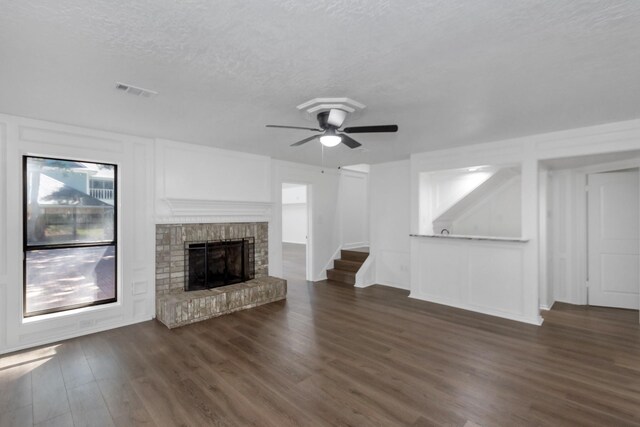 unfurnished living room featuring a textured ceiling, visible vents, dark wood finished floors, a brick fireplace, and a ceiling fan