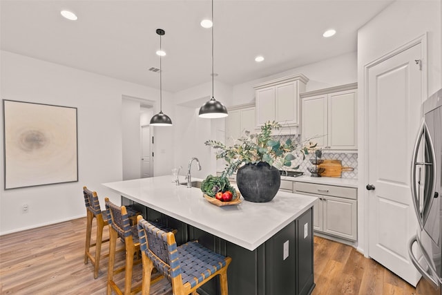 kitchen featuring decorative backsplash, light wood-type flooring, light countertops, stainless steel fridge, and an island with sink