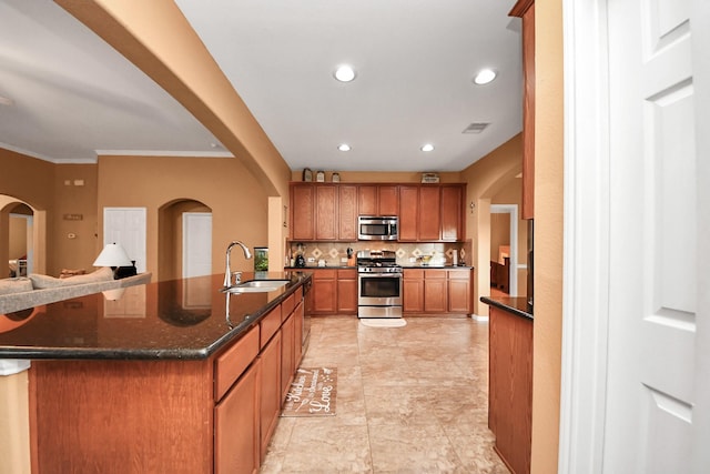 kitchen with brown cabinetry, appliances with stainless steel finishes, tasteful backsplash, a center island with sink, and a sink