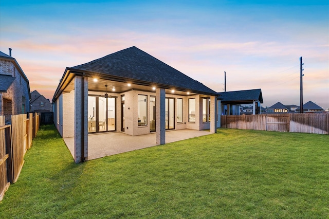 back of property at dusk featuring roof with shingles, a fenced backyard, ceiling fan, a patio area, and a lawn