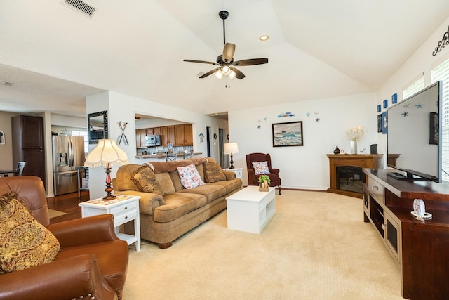 living area with a glass covered fireplace, a ceiling fan, visible vents, vaulted ceiling, and light colored carpet
